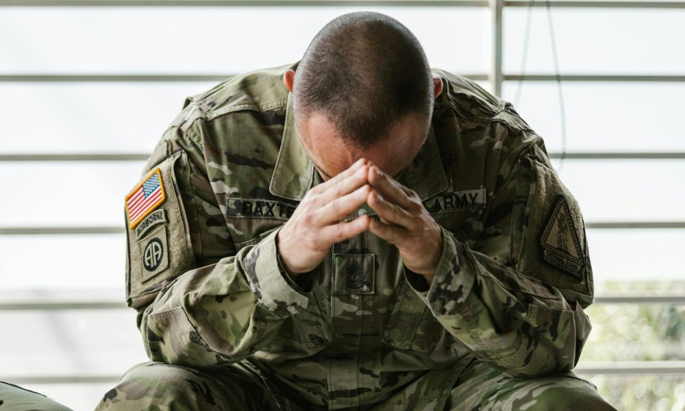 A soldier in camouflage sitting indoors, appearing stressed and overwhelmed.