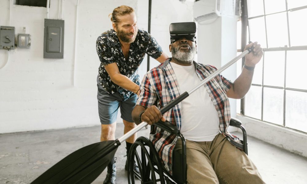 Two men enjoying virtual reality; one is in a wheelchair experiencing a rowing simulation.